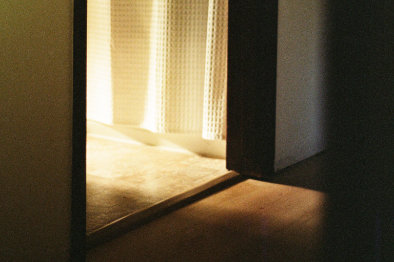 Sunlight streams through a bathroom's doorway into a hallway. A white shower curtain hangs within the bathroom.