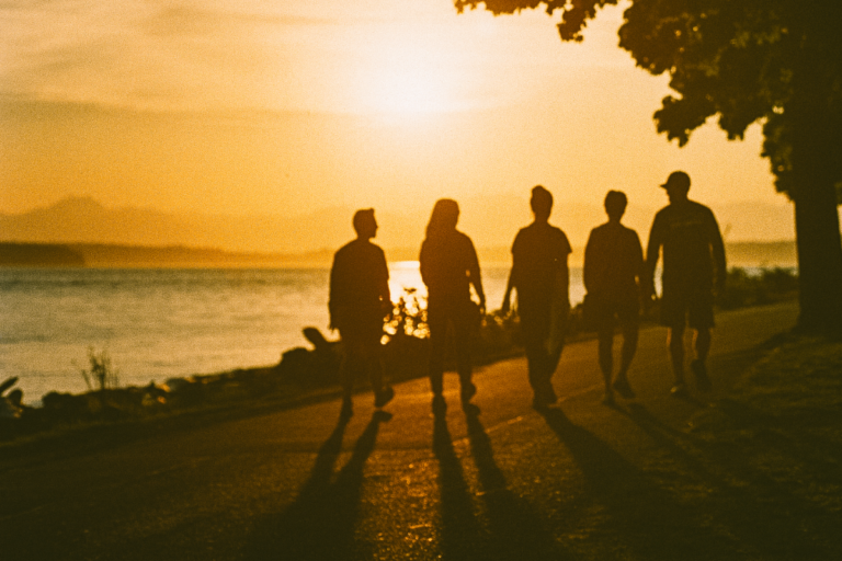 Five people walk on a paved path by a body of water as the sun sets.