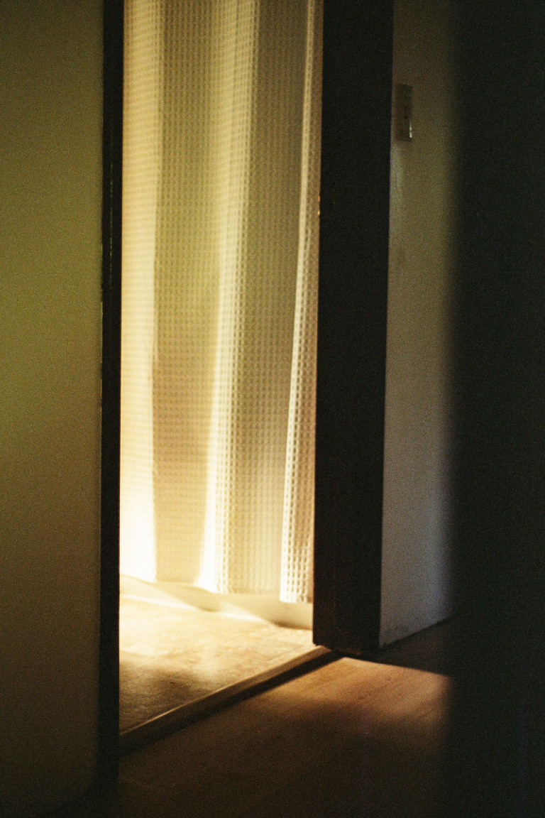 Sunlight streams through a bathroom's doorway into a hallway. A white shower curtain hangs within the bathroom.