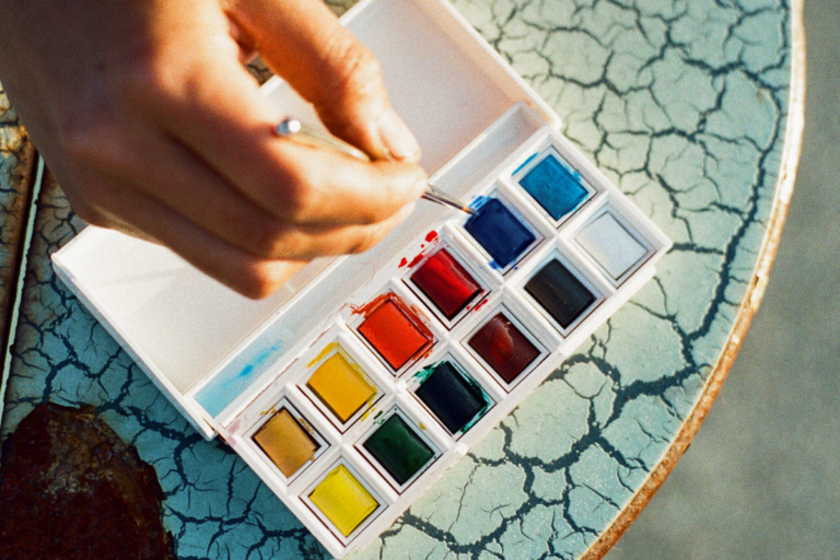 A hand dips a paintbrush into the blue paint of a watercolor palette. The palette sits on a blue crackled table.
