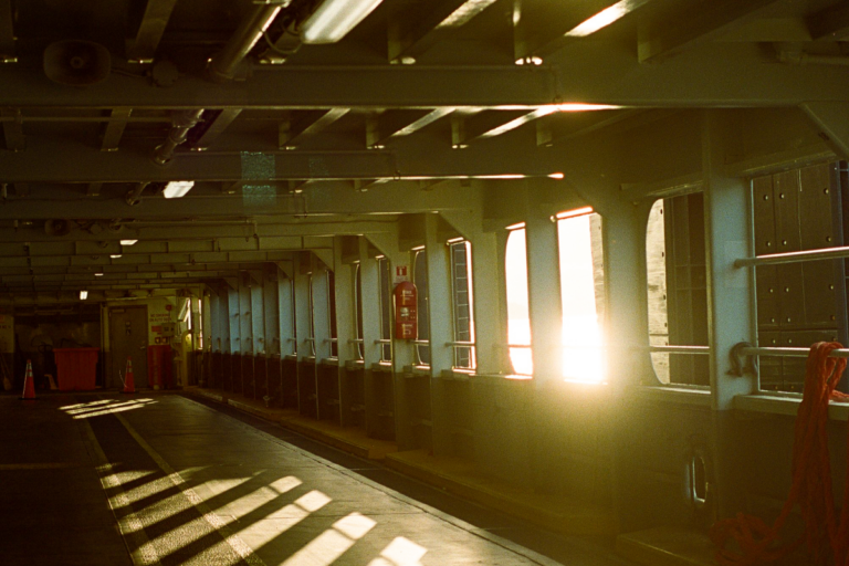 Sunlight streams through openings on the side of a ferry. Shadows created by openings on the side of the ferry are visible on a covered pathway on the ferry.