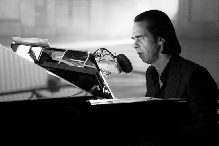 Nick Cave sings while sitting at a piano.