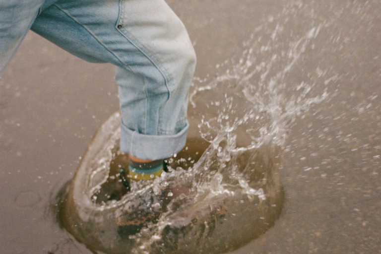 An individual in jeans makes a splash while stepping in water on top of pavement.